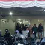Ilustrasi lorong IGD rumah sakit yang padat dengan ranjang pasien dan ambulans bersiaga di luar pada malam hari